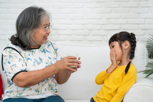 Happy elderly Asian Grandma sits beside her granddaughter and feeds fresh milk from glass for breakfast at home. Concept of a happy family and takes care together, preschool health care photo