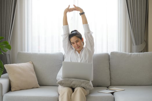 Businesswoman wearing white shirt sitting on sofa with laptop raising her arms above her head stretching to relieve pain after sitting for a long time at work. photo