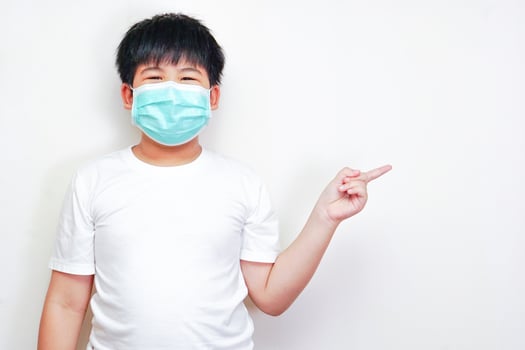 The asian boy in a medical surgical mask, wearing a white t-shirt and pointing his finger to the side. photo