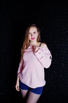 Studio shot of brunette girl at pink sweater with jeans shorts against black brick wall. photo