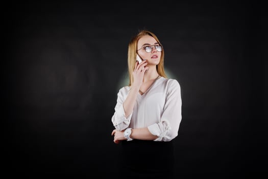 Studio portrait of blonde businesswoman in glasses, white blouse and black skirt speaking at mobile phone against dark background. Successful woman and stylish girl concept. photo
