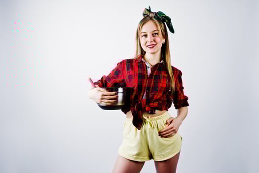 Young funny housewife in checkered shirt and yellow shorts pin up style with saucepan and kitchen spoon isolated on white background. photo