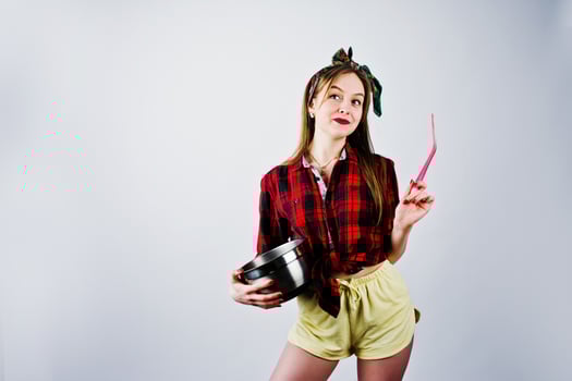 Young funny housewife in checkered shirt and yellow shorts pin up style with saucepan and kitchen spoon isolated on white background. photo