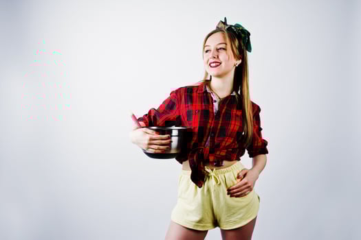 Young funny housewife in checkered shirt and yellow shorts pin up style with saucepan and kitchen spoon isolated on white background. photo