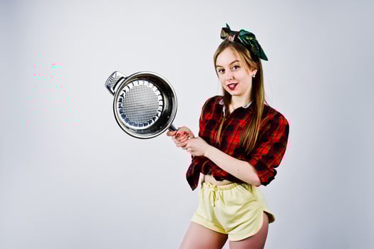 Young funny housewife in checkered shirt and yellow shorts pin up style with frying pan isolated on white background. photo