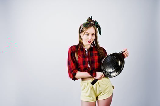 Young funny housewife in checkered shirt and yellow shorts pin up style with frying pan isolated on white background. photo