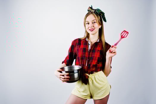 Young funny housewife in checkered shirt and yellow shorts pin up style with saucepan and kitchen spoon isolated on white background. photo