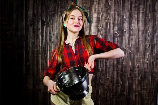 Young funny housewife in checkered shirt and yellow shorts pin up style with kitchen sieve on wooden background. photo