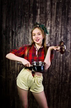 Young funny housewife in checkered shirt and yellow shorts pin up style with saucepan on wooden background. photo