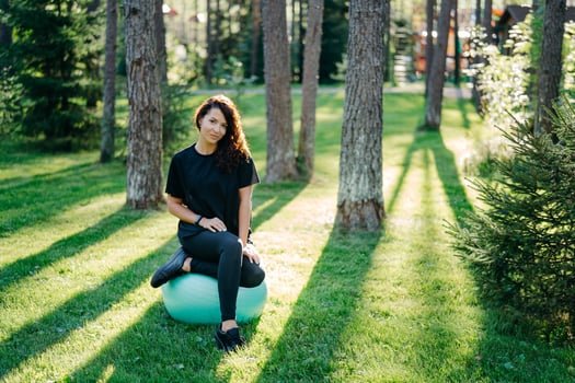 Fit brunette young woman takes rest after doing exercises at fitness ball leads active sporty lifestyle poses in urban environment on green grass against trees with sunshine does gymnastics. photo