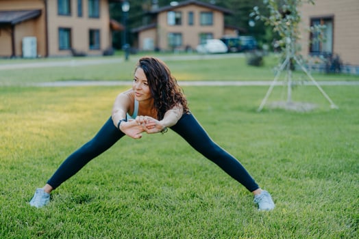 Confident athletic woman demonstrates her flexibility, stretches outdoors, does exercises early in morning on green grass near house leads active lifestyle dressed in active wear stays healthy photo