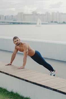 Outdoor shot of determined muscular man warms up before training stands in plank pose has naked torso muscular body enjoys exercising in open air wears active wear has workout to maintain vitality photo