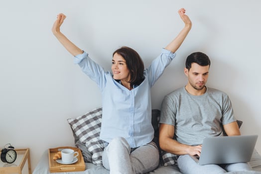 Young married couple spend morning in bed beautiful brunette female stretches and her husband with serious expression uses laptop computer for remote work, dressed casually, being in bedroom photo