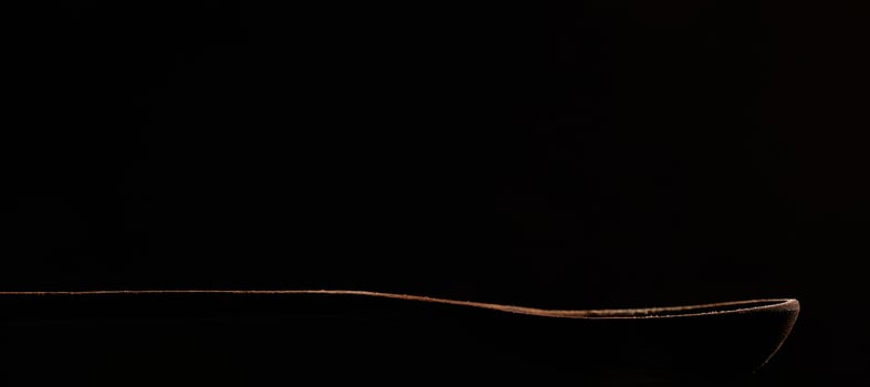 Outline of a wooden brown spoon on a black background. Spoon close-up in the dark. A contour illuminated by light. Photo with copy space.