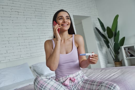 Happy young woman holding pregnancy test and talking on mobile phone while sitting on bed photo