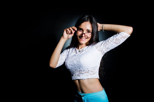 Portrait of an attractive young woman in white top and blue pants posing with her glasses in the dark. photo