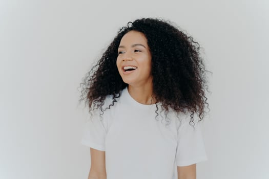 Portrait of beautiful curly adult woman turns gaze aside, has glad expression, smiles and laughs at something funny, dressed in white t shirt, models indoor, feels free, energized and relieved, photo
