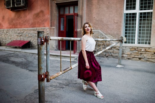 Fashionable and beautiful blonde model girl in stylish red velvet velour skirt, white blouse, hat and handbag, posed against barrier. photo