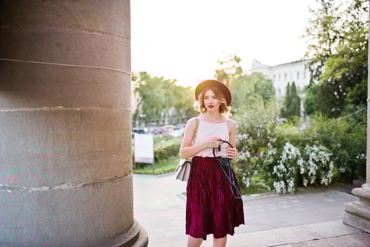 Fashionable and beautiful blonde model girl in stylish red velvet velour skirt, white blouse and hat, posed with phone and earphones against column at sunset. photo