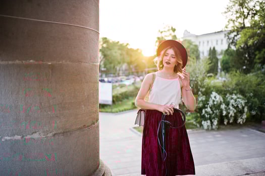 Fashionable and beautiful blonde model girl in stylish red velvet velour skirt, white blouse and hat, posed with phone and earphones against column at sunset. photo