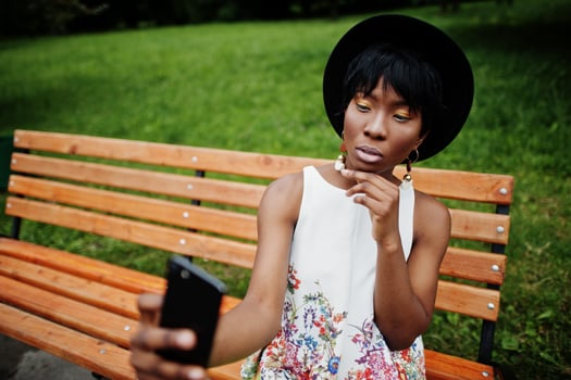 Amazing african american model woman in green pants and black hat posed on bench at park and making selfie on mobile phone. photo