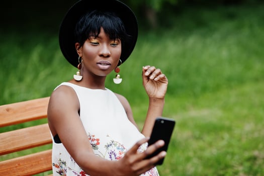 Amazing african american model woman in green pants and black hat posed on bench at park and making selfie on mobile phone. photo