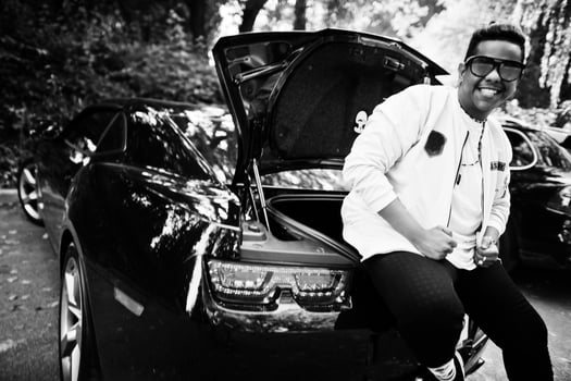 Stylish hipster arab man guy in sunglasses posed outdoor in street and sitting on the trunk his black muscle car. Rich black man. photo