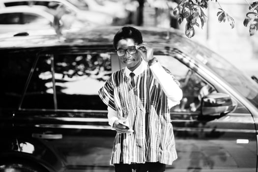 African businessman in traditional clothes and glasses with mobile phone against black car suv. Rich africans people. photo