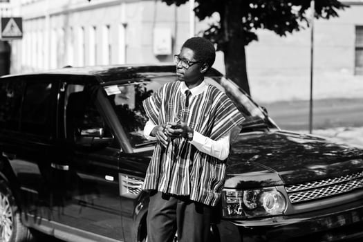 African businessman in traditional clothes and glasses with mobile phone against black car suv. Rich africans people. photo
