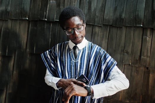 African businessman in traditional clothes and glasses with mobile phone at hand looking at his watches against wooden wall. photo