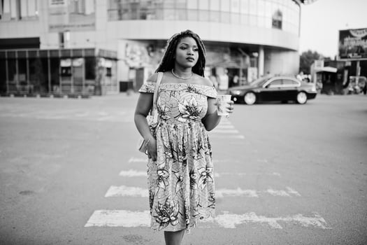 Cute small height african american girl with dreadlocks, wear at coloured yellow dress, walking at crosswalk against trade center with cup of coffee at hand. Black and white. photo