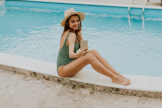 Young woman sitting by the swimming pool and taking selfie photo with  mobile phone in the house backyard