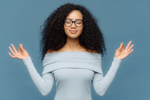 Relaxed beautiful woman with Afro hairstyle, feels relieved, shows zen gesture, keeps hands in okay gesture, controls emotions, gets in control with herself has eyes closed isolated on blue background photo