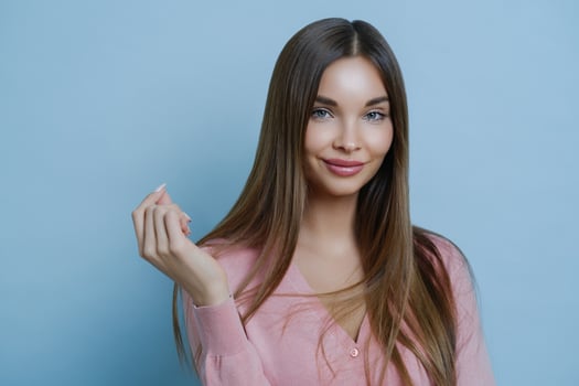 Beautiful young European lady with no blemishes and makeup, has cute look, keeps hand raised, feels delighted, poses against blue background, wears rosy jumper, going to participate in fashion show photo