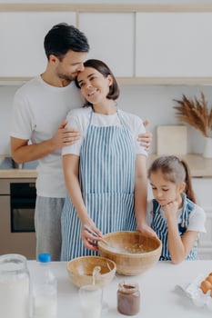 Family cook meal at home. Husband and wife embrace with love, small girl poses near, prepare delicious dinner, use milk, chocolate, eggs, make homemade pastry, pose against kitchen interior. photo