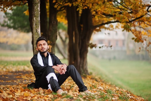 Indian stylish man in black traditional clothes with white scarf posed outdoor against yellow autumn leaves tree. photo