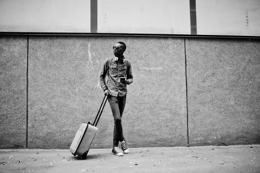 African american man in checkered shirt, sunglasses and earphones with suitcase. Black man traveler against wall holding mobile phone. photo