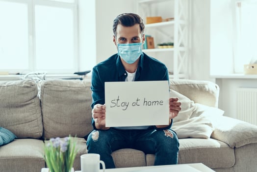 Young man in protective face mask showing stay at home saying written on the paper while sitting on sofa photo