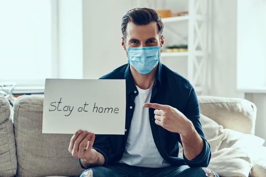 Handsome young man in protective face mask showing stay at home saying written on the paper while sitting on sofa photo