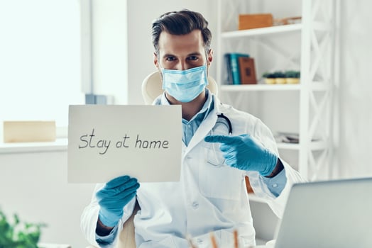 Young man in white lab coat and protective mask showing stay at home saying written on the paper while sitting indoors photo