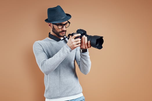 Charming young man in casual wear adjusting photo camera while standing against brown background
