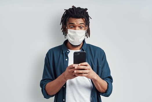 Surprised young African man wearing medical face mask and using smart phone while standing against grey background photo
