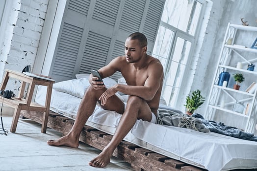 Checking messages. Sleepy young African man using his smart phone while sitting on the bed at home photo