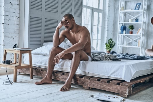 Too early to wake up. Sleepy young African man covering eyes with hand while sitting on the bed at home photo