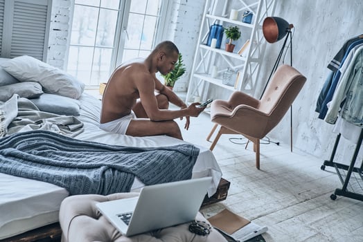 Chatting with girlfriend. Sleepy young African man using his smart phone while sitting on the bed at home photo