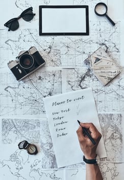 Places I want to visit. Close up top view of man writing down checklist with sunglasses, photo camera, compass, magnifying glass and passport lying on map around