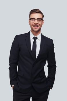 Perfect in his style. Handsome young man in full suit keeping hands in pockets and looking at camera with smile while standing against grey background photo