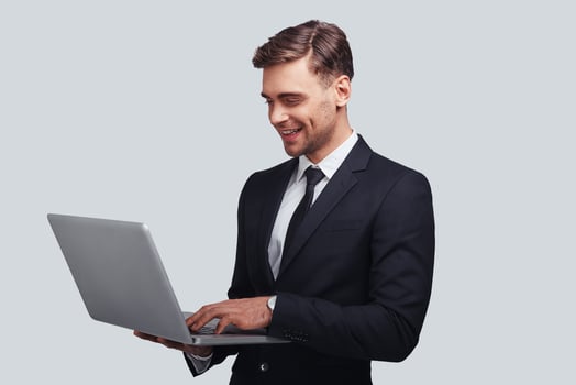 Ready to work hard. Good looking young man in full suit using laptop and smiling while standing against grey background photo