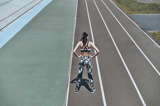 Training to become the best. Top rear view of young woman in sports clothing keeping hands on hips while standing on the running track photo