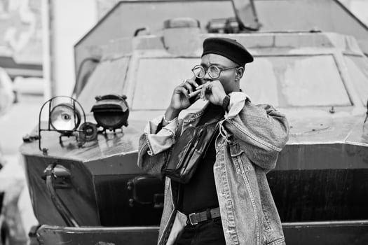 African american man in jeans jacket, beret and eyeglasses, with cigar posed against btr military armored vehicle, and speaking on mobile phone. photo